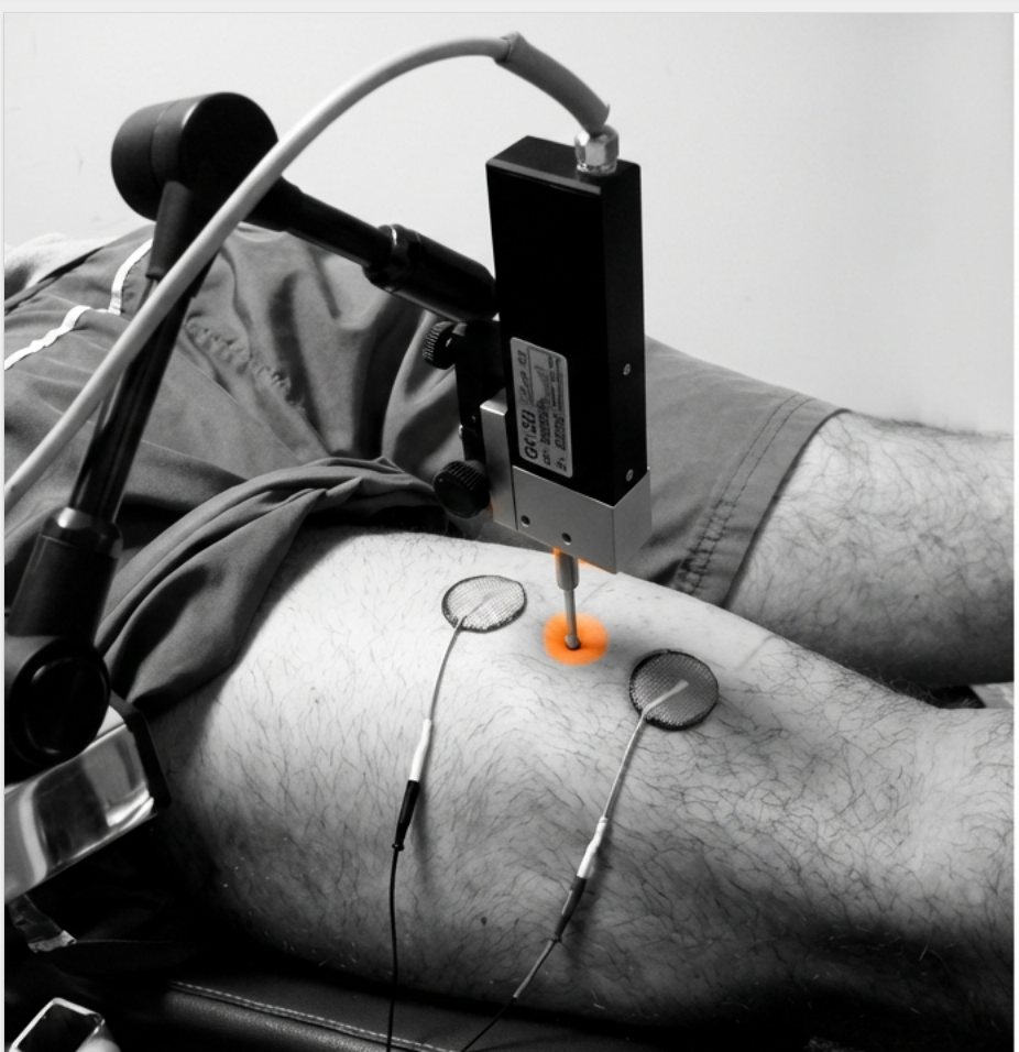 The TMG sensor touching the skin over a thigh muscle, between two electrodes. The sensor tip glows orange.